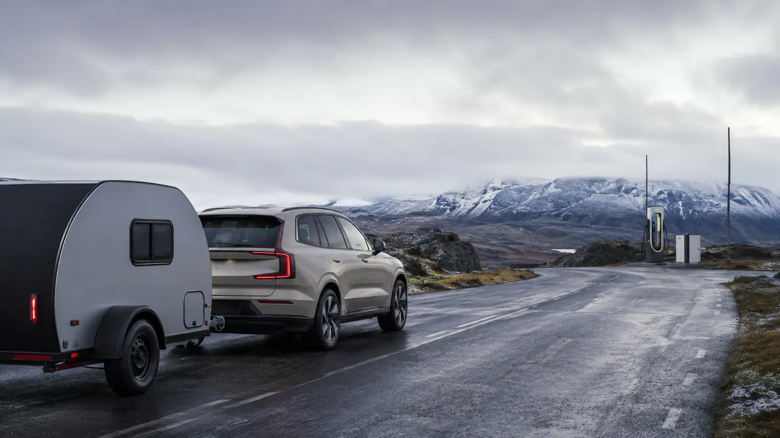 Luxury electric SUV towing a compact trailer on a cold mountain road near a charger